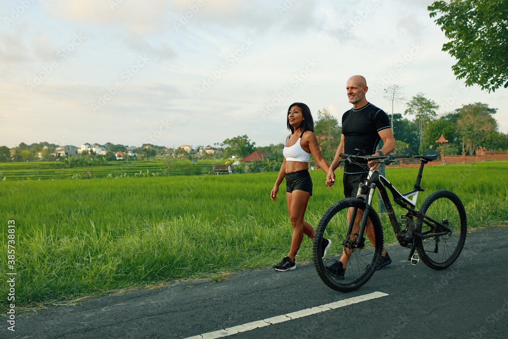 Sportive Couple Walking Bike On Road. Asian Woman And Caucasian Man Walking By Hands With Bicycles And Talking Against Green Tropical Landscape. Sportive Couple Walking Bike On Road. Asian Woman And Caucasian Man Walking By Hands With Bicycles And Talking Against Green Tropical Landscape.
