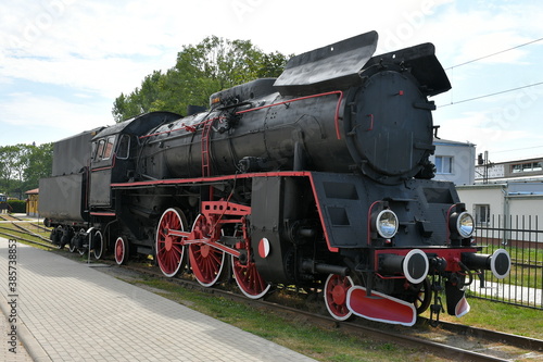 Close up on a vintage train with a black metal body and red and white wheels standing on a side track surrounded with some trees and with a train station visible in the background in Poland