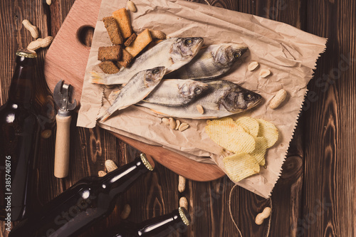 Brown glass bottles of beer and dried fish with chips, nuts, crackers on paper on a wooden background.