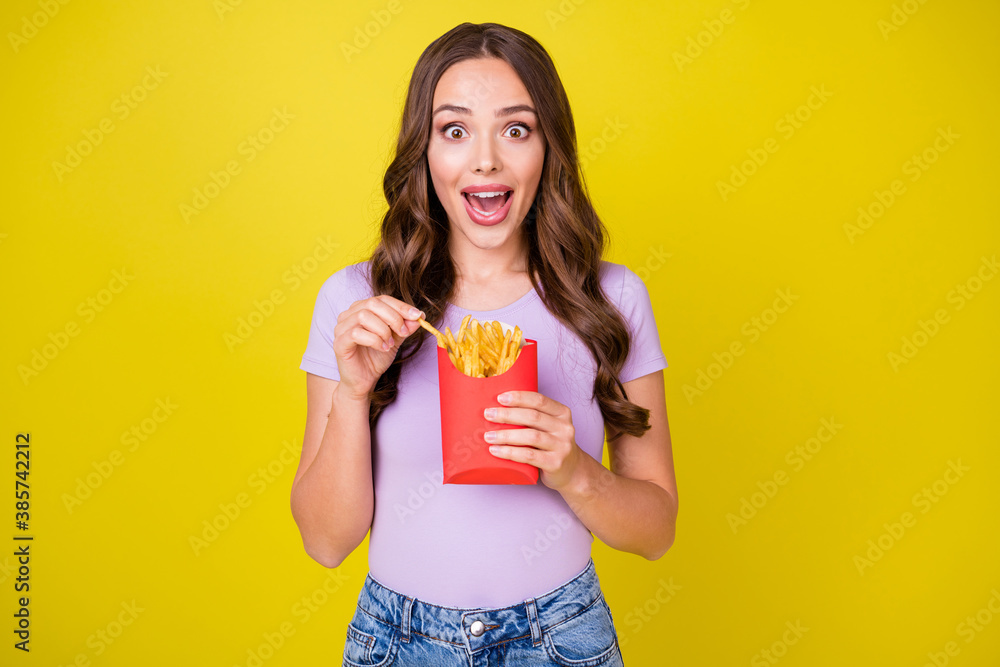 Portrait of attractive amazed hungry cheerful wavy-haired girl eating french fries isolated over bright yellow color background - obrazy, fototapety, plakaty 