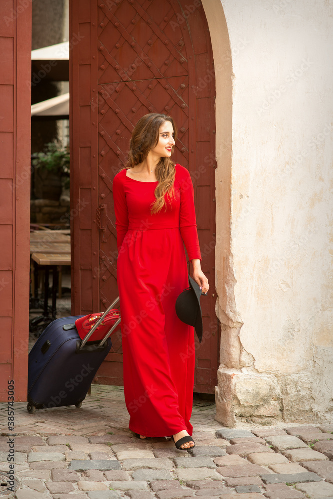 Young caucasian woman comes out of the ancient door with baggage wearing long red dress on the old city street
