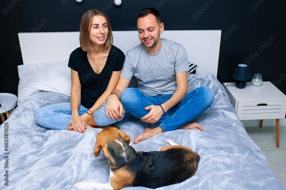 Young couple sitting on a bed in the bedroom, hugging. Near them is their dog Young couple sitting on a bed in the bedroom, hugging. Near them is their dog