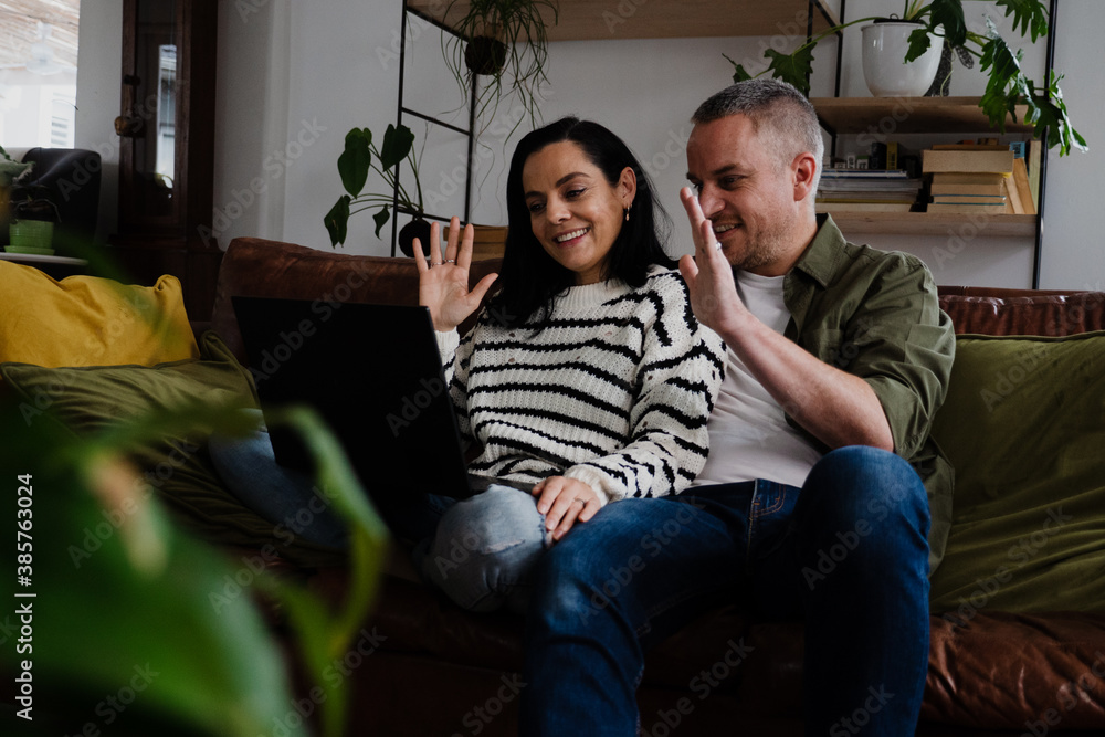 Happy couple smiling and waving while on video call with overseas family using laptop sitting on sofa in lounge. Happy couple smiling and waving while on video call with overseas family using laptop sitting on sofa in lounge.