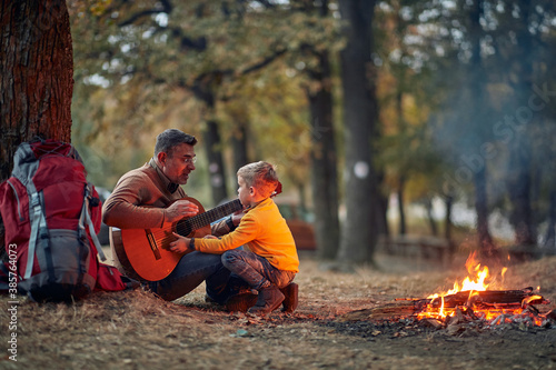 Grandfather and grandson playing a guitar around campfire in the forest
