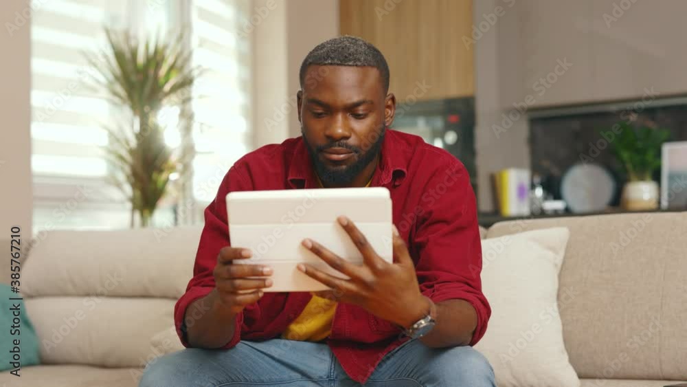 Serious african american young man using digital tablet computer at home sitting on sofa. Handsome guy. Technology thinking black apartment online indoor. Slow motion Serious african american young man using digital tablet computer at home sitting on sofa. Handsome guy. Technology thinking black apartment online indoor. Slow motion