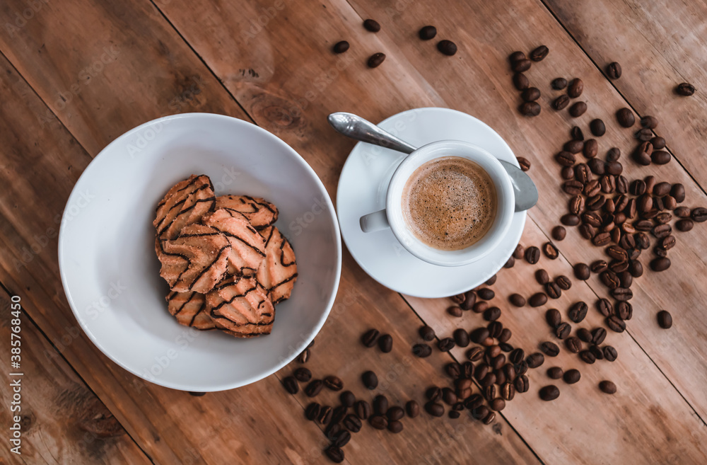 cup of espresso accompanied by cookies with chocolate on a white plate, coffee beans on a rustic wooden background, top view