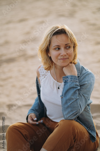beautiful young caucasian smiling woman wearing white blouse and denim jacket stitting on sand at the beach before sunset, touching hair and looking aside. Image with selective focus
