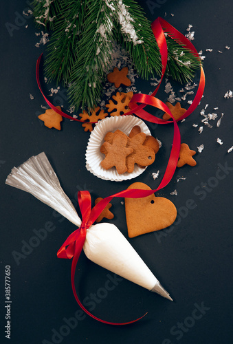 Christmas gingerbread cookies with fir festive decoration with snow and pastry bag with icing on black background, view from above. Holiday food, homemade baking, Christmas and New Year traditions.