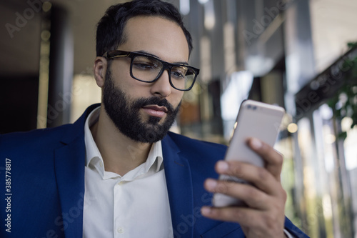 Young pensive middle eastern man using mobile phone, reading news, working online in modern office. Portrait of handsome arabic businessman wearing stylish eyeglasses, looking at digital screen