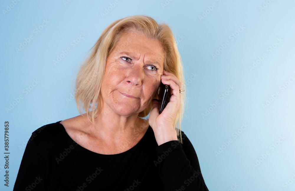 Studio portrait of 60+ woman with blonde page looking concerned while making a phone call on light blue background. Studio portrait of 60+ woman with blonde page looking concerned while making a phone call on light blue background.