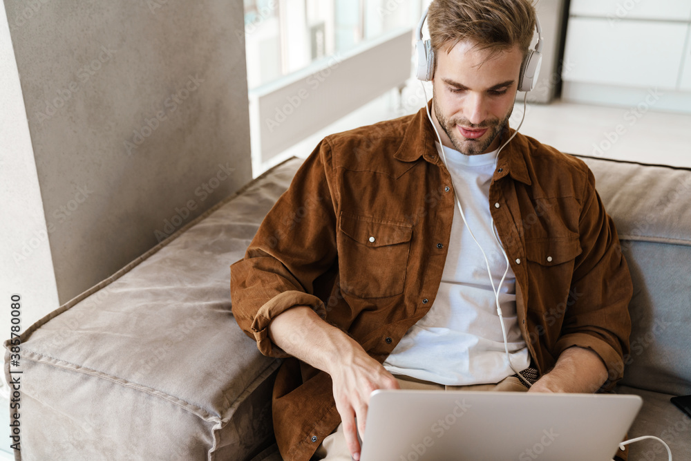 Fototapeta premium Handsome focused guy using headphones and laptop while sitting on sofa