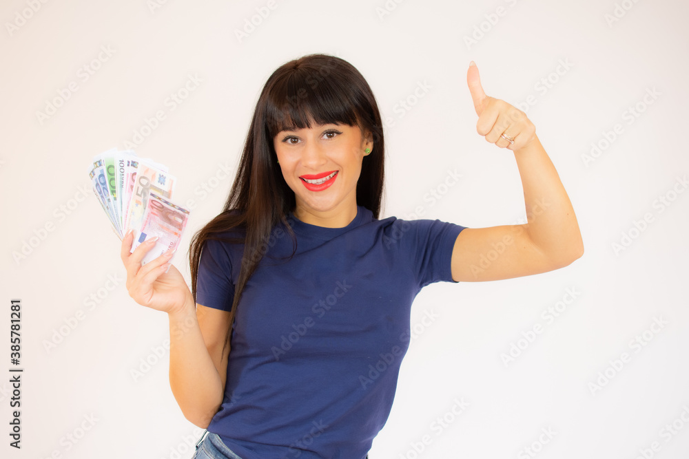 Obraz premium Portrait of a smiling young girl showing bunch of money banknotes and giving thumbs up isolated over white background