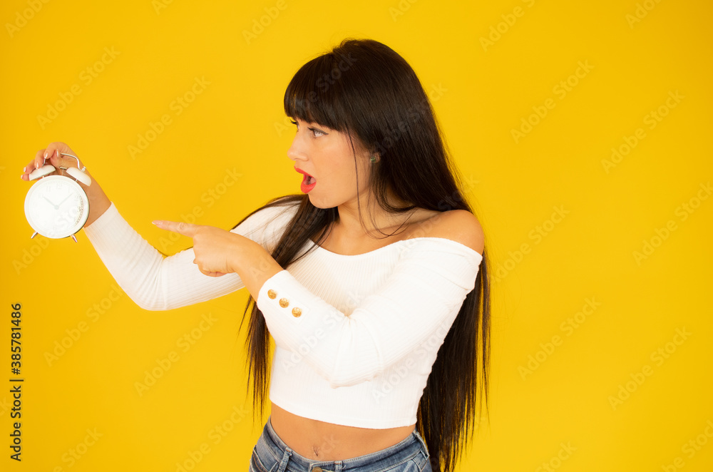 Worried woman in white pullover pointing at the alarm clock, isolated on yellow Worried woman in white pullover pointing at the alarm clock, isolated on yellow