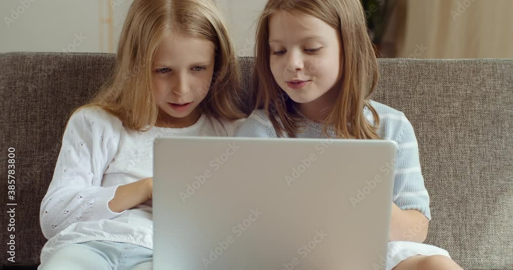 Front view of two little schoolgirls friends sitting together on couch at home in front of laptop screen watching cartoon movie online, laughing talking, sisters kids having fun spending time together Front view of two little schoolgirls friends sitting together on couch at home in front of laptop screen watching cartoon movie online, laughing talking, sisters kids having fun spending time together