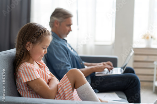 No Time For Child. Grey-haired dad busy with laptop, working online at home, sad bored offended daughter sitting near by.