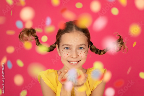 Fancy child with pigtails blowing confetti against pink background