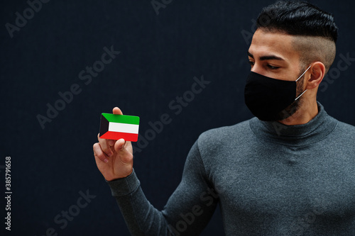 Middle eastern man in gray turtleneck and black face protect mask show Kuwait flag isolated background. photo