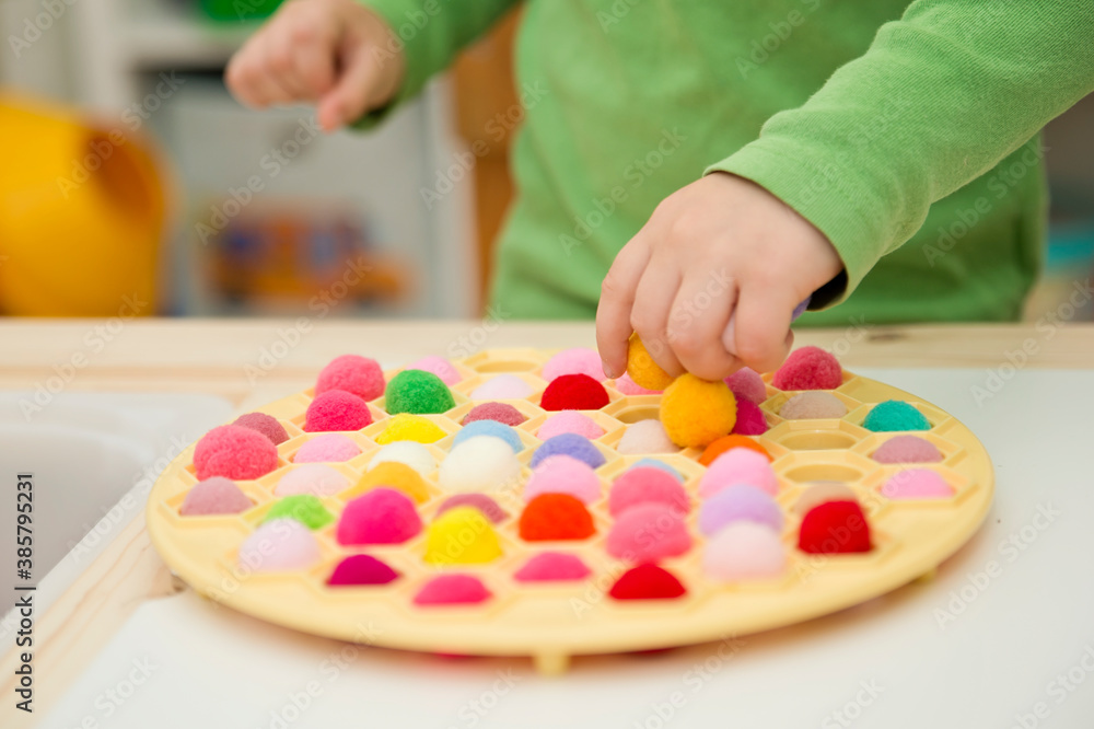2-year child playing with Russian Dumpling Maker by inserting pompoms in holes. colored yarn balls in plastic basket 2-year child playing with Russian Dumpling Maker by inserting pompoms in holes. colored yarn balls in plastic basket