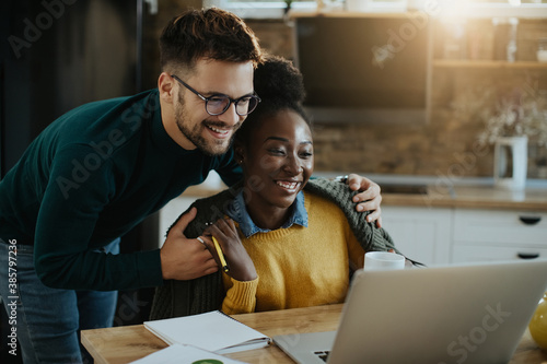 Happy mixed-race couple reading an e-mail on laptop at home.