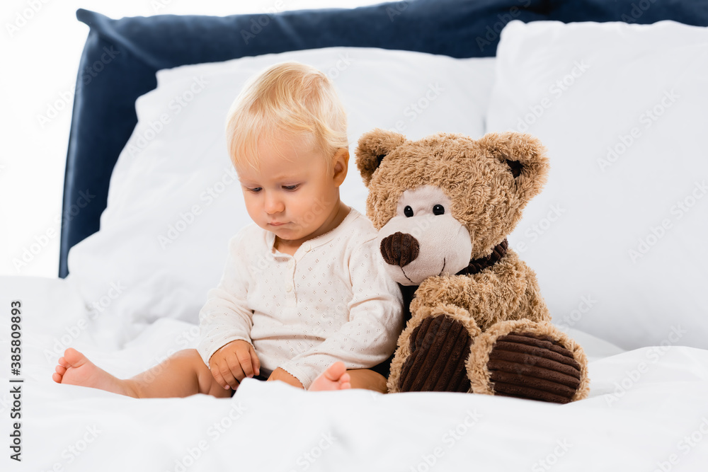 Selective focus of toddler sitting near soft toy on bed on white background