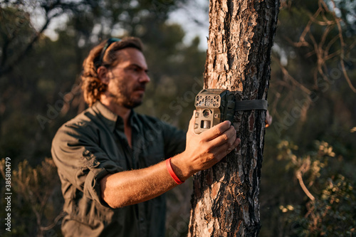 Mature man holding trail camera on tree trunk while standing in forest