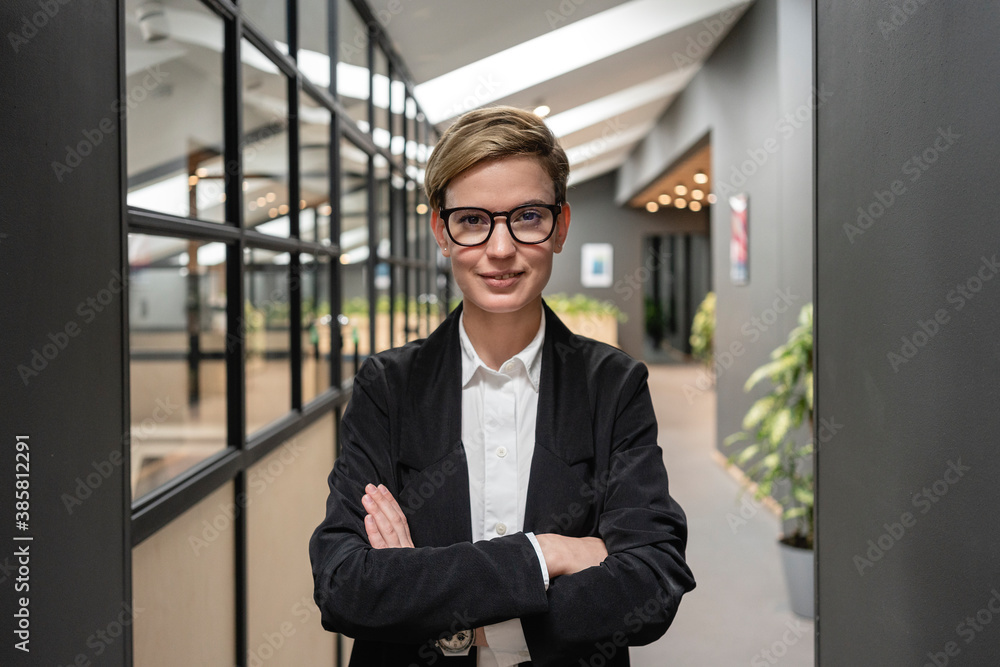 Smiling beautiful female professional standing with arms crossed at illuminated coworking office corridor Smiling beautiful female professional standing with arms crossed at illuminated coworking office corridor