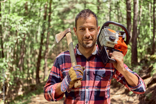 Smiling lumberjack standing with axe and chainsaw in forest photo