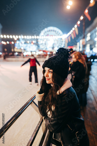 Cute girl in a hat and winter clothes stands near rink on the background of the Christmas street with lights and a Ferris wheel, looking to side.Lady posing against the backdrop of the christmas fair.