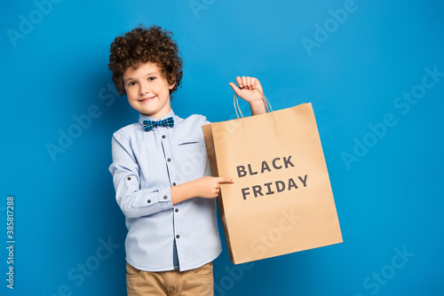 joyful and curly boy pointing with finger at shopping bag with black friday lettering on blue © LIGHTFIELD STUDIOS
