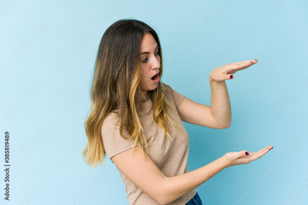 Young caucasian woman isolated on blue background shocked and amazed holding a copy space between hands. Young caucasian woman isolated on blue background shocked and amazed holding a copy space between hands.