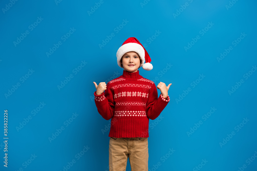 Obraz premium joyful kid in santa hat and red sweater showing thumbs up while looking at camera on blue