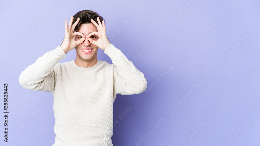 Young caucasian man isolated on purple background showing okay sign over eyes Young caucasian man isolated on purple background showing okay sign over eyes