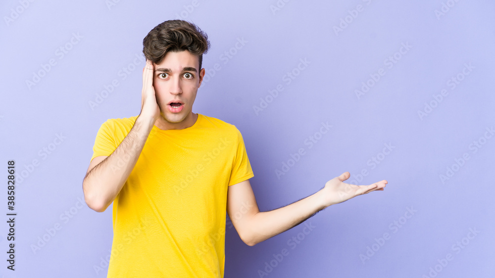 Fototapeta premium Young caucasian man isolated on purple background impressed holding copy space on palm.