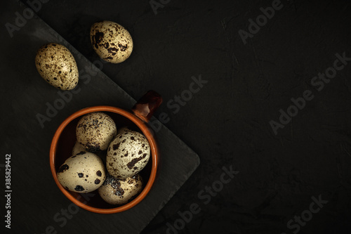 raw quail eggs in a brown clay cup and on a black stone plate on a textured dark concrete surface. moody artistic mockup with copy space. top view
