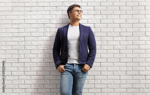 Smiling young man in jeans and suit leaning on a white brick wall