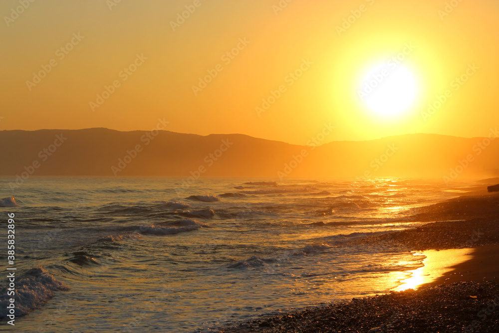 Big yellow sun setting over the sea. Waves hit the pebble beach. Mountains in the background