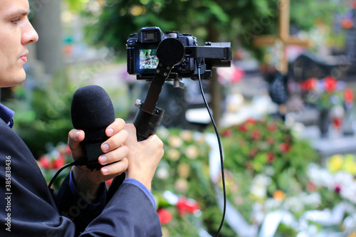 Elegant man in suit is recording grave with cross full of flowers. Funeral live streaming on cementary. Cameraman handing a microphone and camera on a gimbal.