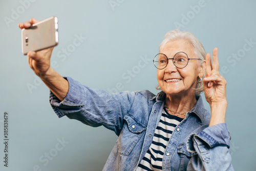 Positive smiling elderly woman wearing casual clothes and glasses poses for selfie holding smartphone in one hand and making peace or victory sign with another. Active old age concept.