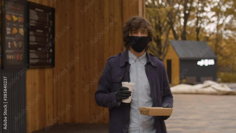 Young handsome man in a protective mask and gloves comes out of a fast food restaurant with a food box and a plastic cup in his hands. Autumn day in the city Young handsome man in a protective mask and gloves comes out of a fast food restaurant with a food box and a plastic cup in his hands. Autumn day in the city