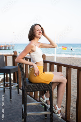A girl in a yellow skirt and white top sits at a chair on the summer terrace of a cafe. On vacation in Turkey. Beach and sea in the background. Smile. Sunny day. Vocation. Lifestyle. Rest. Relax. © Катерина Бухвалова