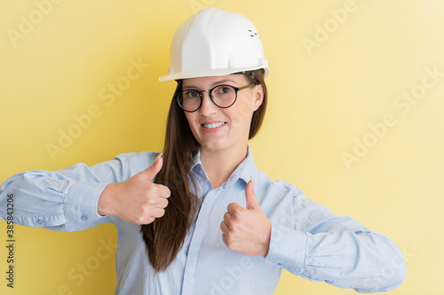 Portrait of smiling beautiful tatar woman engineer in construction helmet and glasses showing thumbs up isolated on yellow background