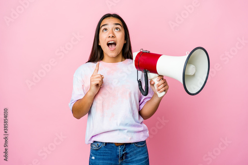 Young asian woman holding a megaphone isolated on pink background pointing upside with opened mouth.