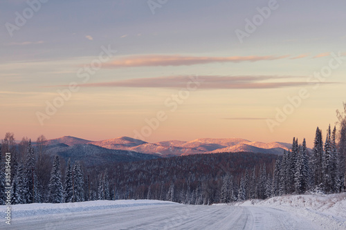 The road in the winter mountains in the background. Winter valley panorama with forest and mountain peaks in the background at sunrise. First rays of the sun in the morning
