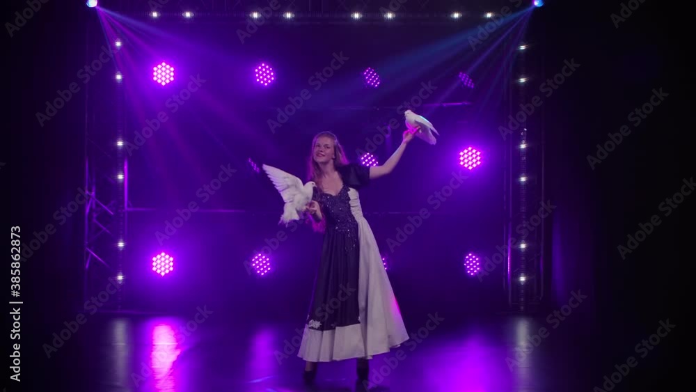 A young woman trainer in a long dress performs in the studio with her trained white doves. Birds sit on their arms and flap their wings in studio light. Festive performance of pigeons. Slow motion. A young woman trainer in a long dress performs in the studio with her trained white doves. Birds sit on their arms and flap their wings in studio light. Festive performance of pigeons. Slow motion.