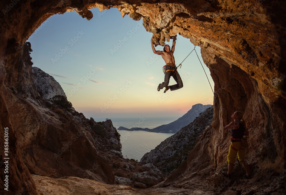 Rock climbing at sunset. Young man climbing route in cave, female partner belaying him.