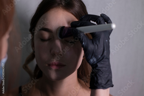 the process of applying makeup to a beautiful brunette by a master in a medical mask and black gloves. beauty  modeling and cosmetics concept.