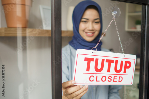 cheerful mature woman in closed sign and smiling while standing against front door office studio. Tutup in english mean close