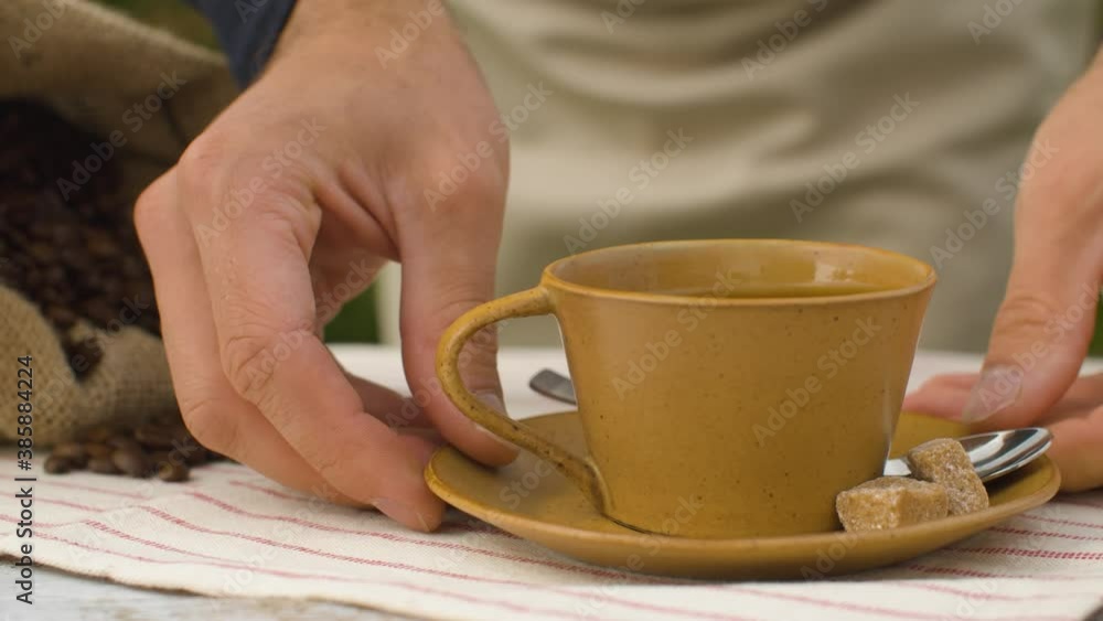 Close-up of coffee beans in a burlap jute sack and cup of coffee on a table outdoors. Hands placing a cup. Dolly shot Close-up of coffee beans in a burlap jute sack and cup of coffee on a table outdoors. Hands placing a cup. Dolly shot