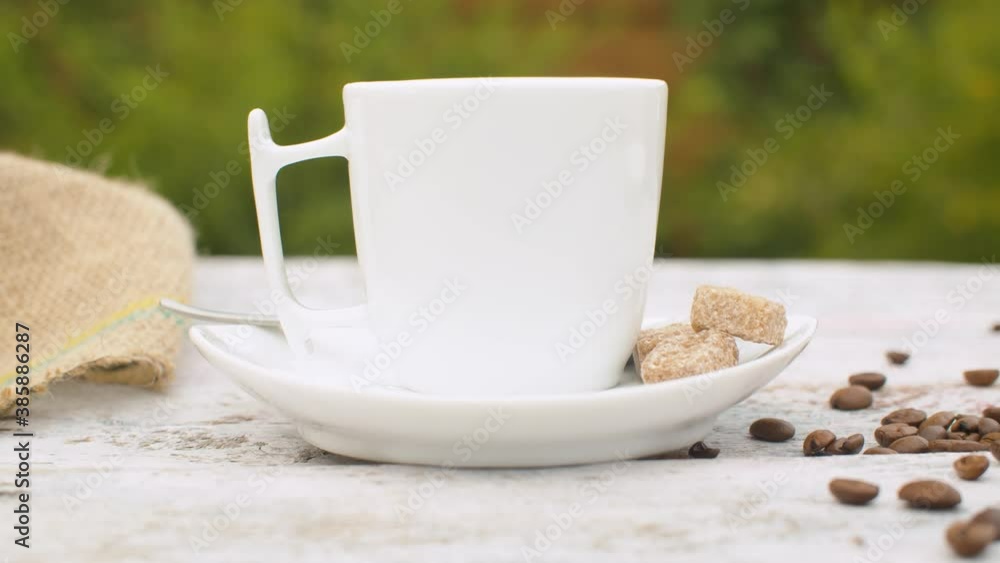 Close-up of cup of hot coffee, raw sugar and coffee beans on a table in the garden. Steam rises from the cup. Dolly shot Close-up of cup of hot coffee, raw sugar and coffee beans on a table in the garden. Steam rises from the cup. Dolly shot