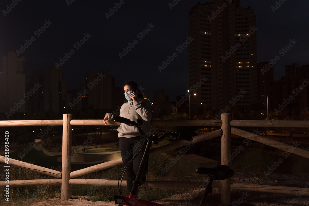 Young woman with protective face mask and casual clothes calling on her smart phone at night.